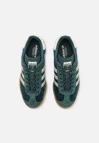 Zapatillas de deporte verde de ante con acentos de tres rayas blancas, cordones planos y suela de goma. Presenta un logo de Adidas en la lengüeta.