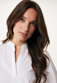 Blusa bianca con colletto arricciato e placca. Tessuto leggero e liscio con un leggero lucido. La modella ha lunghi capelli ricci castani.