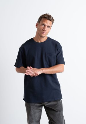 Tummansininen puuvillainen t-paita, jossa on pyöreä kaula-aukko, lyhyet hihat ja rintatasku. Siinä on rento istuvuus ja pehmeä pinta.