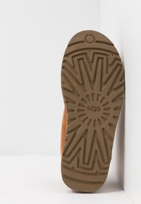Semelle marron d'une botte UGG avec un motif rayons de soleil texturé et un motif de bande de roulement géométrique sur fond blanc dans un coin.