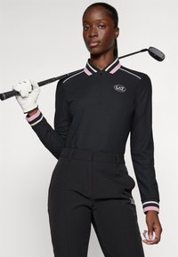 Svart golfpolo med krage och logotyp i rosa ränder, tillsammans med svarta byxor. Golfaren håller en klubba och en golfboll.
