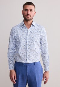 Muž s krátkymi vlasmi a bradou, oblečený v modrej kvetinovej košeli na gombíky a modrých nohaviciach, stojaci pred jednofarebným svetlým pozadím.