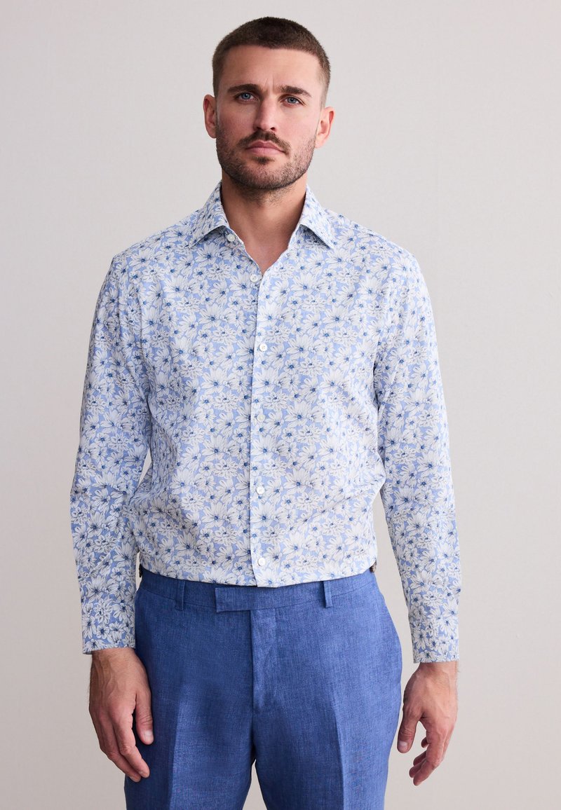 Muž s krátkymi vlasmi a bradou, oblečený v modrej kvetinovej košeli na gombíky a modrých nohaviciach, stojaci pred jednofarebným svetlým pozadím.