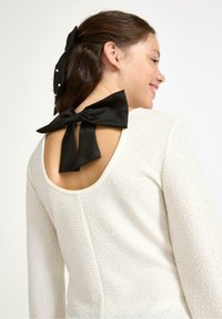 Biała, teksturowana bluzka z wykrojem na plecach i dużą czarną satynową kokardą. Projekt ma długie rękawy i gładki dekolt.