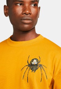 Sweatshirt galben din bumbac, cu un design imprimat al unei păianjeni negri și maro pe față, având detalii complicate pe corpul și picioarele păianjenului.