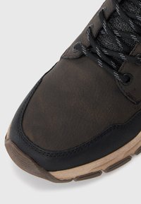 Punta di scarpa da trekking in pelle marrone con bordi neri, lacci neri e grigi a macchie e suola in gomma beige su uno sfondo bianco.