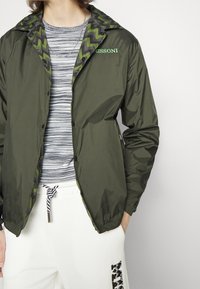 Rompevientos de color verde oliva con un interior de patrón en zigzag negro y verde. Cuenta con botones a presión, un cuello y un logotipo bordado en el pecho.