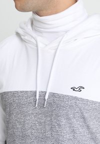 Hette-tröja med vit överdel och strukturerad grå botten, med en logotyp-accent och vita dragsnören. Slät bomullsblandning.