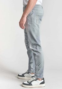 Jasnoniebieskie, zniszczone dżinsy o slim fit, z prostymi nogawkami, pięcioma kieszeniami i subtelnymi efektami blaknięcia. Połączone z czarno-białymi sneakersami.