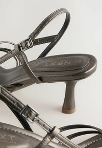 Šedé metalické sandály na podpatku s několika pásky. Disponují elegantním povrchem, nastavitelnými sponami a strukturovaným podpatkem pro zvýšenou přilnavost.
