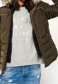 Chaqueta acolchada verde oliva con capucha forrada de piel, bolsillos con cremallera y detalles plateados, superpuesta sobre una sudadera gris con texto impreso.