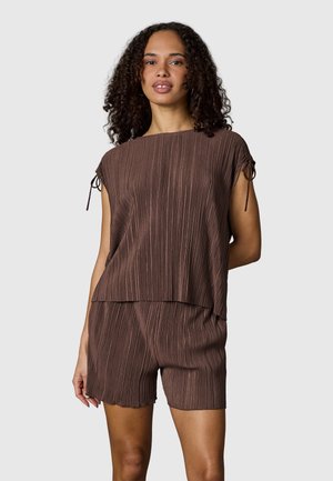 Femme aux cheveux bouclés portant un haut marron plissé à manches courtes et un short plissé assorti, debout devant un fond uni gris clair.