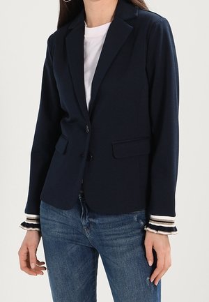 Frau trägt einen marineblauen Blazer über einem weißen Hemd und blauen Jeans, mit gestreiften Manschetten sichtbar unter den Blazerärmeln.