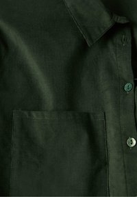 Camicia in corduroy verde scuro con colletto, tasca sul petto e chiusura con bottoni. La texture è a coste con una finitura morbida.