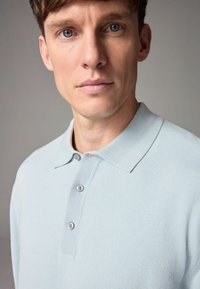 Svetlomodrá pletená polo košeľa so štruktúrovaným golierom, s tromi gombíkmi vpředu a jemnou, hladkou textúrou.
