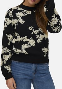 Pull noir avec broderie florale crème, encolure ronde et manches longues ; associé à un jean en denim bleu. Texture douce et design ajusté.