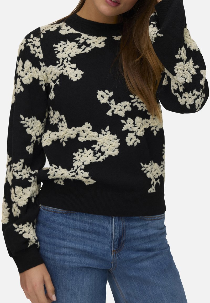 Pull noir avec broderie florale crème, encolure ronde et manches longues ; associé à un jean en denim bleu. Texture douce et design ajusté.