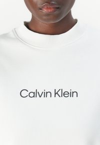 Personne portant un sweat-shirt blanc à col rond avec "Calvin Klein" imprimé en noir sur la poitrine, montrant le bas du visage et le cou.