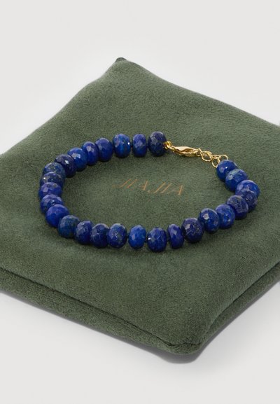 Pulsera de lapislázuli con cuentas redondas facetadas en un azul profundo, asegurada con un cierre de tono dorado, reposando sobre una superficie de exhibición texturizada verde.