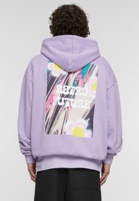 Vijolična prevelika kapuca z velikim grafičnim motivom na hrbtni strani, ki vključuje besedilo "RETRO FUTURE", cvetlične vzorce in abstraktne spiralne vzorce.