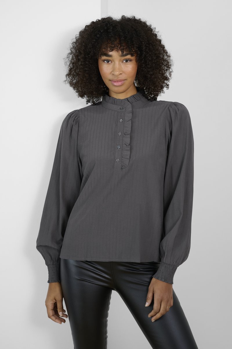 Graue Bluse mit hohem Kragen, gerafftem Vorderblende und langen Puffärmeln. Kombiniert mit einer eleganten schwarzen Kunstlederhose.