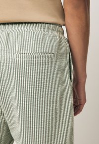 Pantalones de rayas verdes y blancas con tela texturizada, que cuentan con un bolsillo trasero y una cinturilla elástica, mostrando un primer plano del lado derecho.