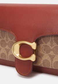 Lähikuva ruskeasta nahasta ja kuvioidusta kankaasta valmistetusta lompakosta, jossa on kultainen metallinen solki, joka on muotoiltu tyylitellyksi "C":ksi valkoista taustaa vasten.