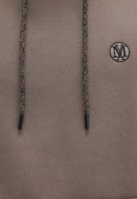 Beige hoodie with black braided drawstrings, featuring black end caps and a circular logo on the chest in dark embroidery.