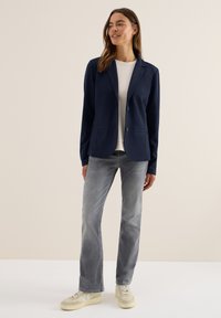 Marinblå blazer med två knappar, slagkrage och framfickor, kombinerad med en ljus tröja med rund hals och grå raka jeans. Mönstrade sneakers.