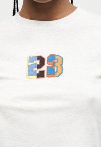 Άτομο που φοράει λευκό T-shirt με κεντημένο αριθμό "23" σε πολύχρωμα τετράγωνα στο κέντρο του στήθους.