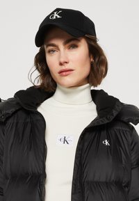 Black puffer jacket with a zip, featuring a white Calvin Klein logo on the chest. Accented by a white ribbed turtleneck and black baseball cap.