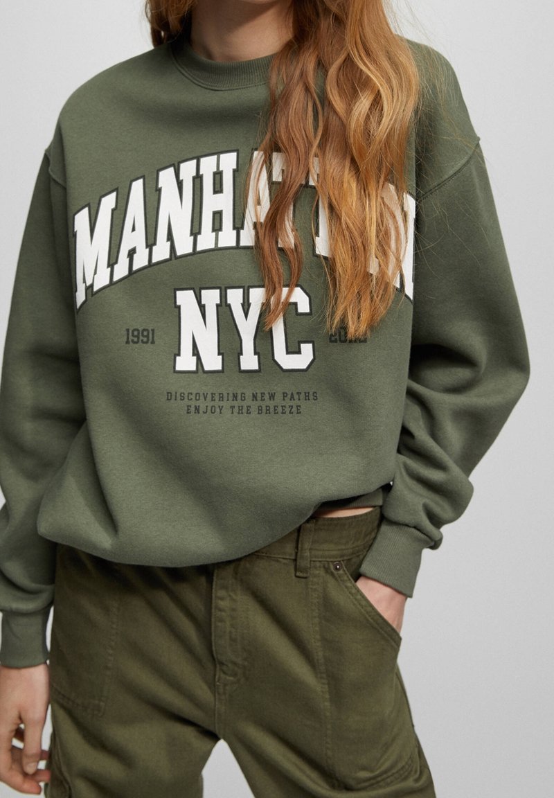 Personne portant un sweat-shirt vert olive avec le texte "Manhattan NYC" et un pantalon vert olive, main dans la poche, cheveux longs ondulés rougeâtres visibles.