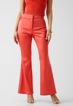 Frau trägt korallenfarbene Schlag-Satin-Hose, ein passendes ärmelloses, strukturiertes Oberteil und goldene, Riemchen-High-Heel-Sandalen, steht vor weißem Hintergrund.