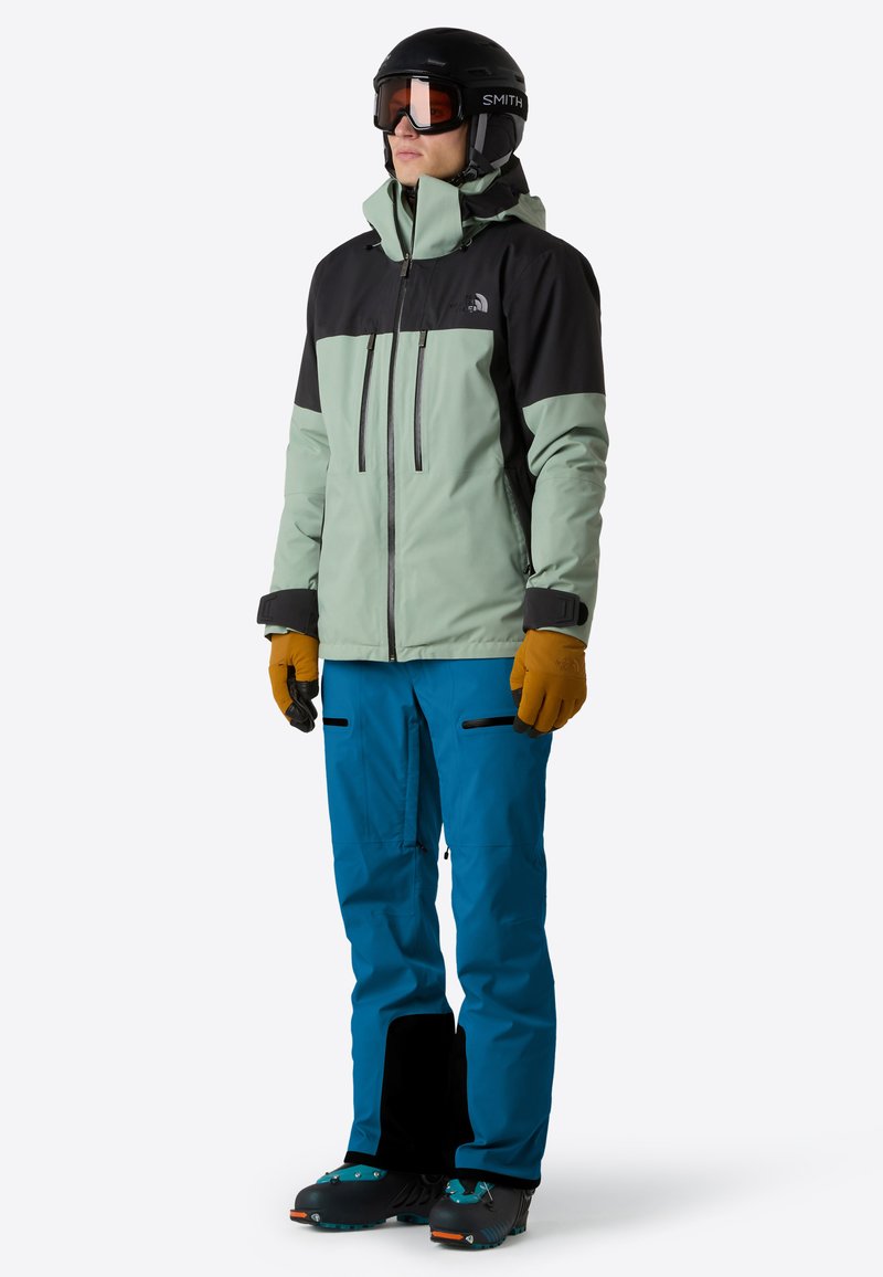 Persona vestita con giacca nera e verde menta, pantaloni da neve blu, casco da sci e occhiali, e guanti marroni, in piedi su uno sfondo bianco.