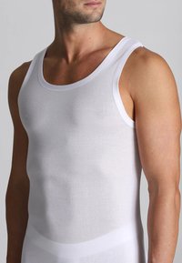 Uomo che indossa una canottiera bianca semplice e aderente, che mostra la parte superiore del corpo e parte del volto su uno sfondo neutro.