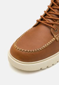 Hnědé kožené boty se šitým špičkovým krytem a kontrastní béžovou gumovou podrážkou. Mají hnědé tkaničky a texturované akcenty podél okraje.
