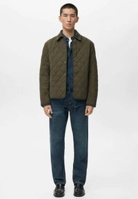 Gequilted olijfgroene jacket met drukknoopsluiting, gedragen over een spijkerhemd. Gecombineerd met donkerblauwe rechte jeans en zwarte schoenen.