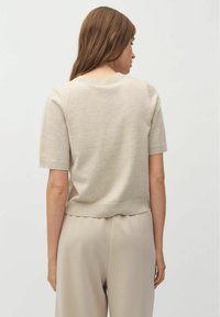 Kurzärmliges, beiges Strickoberteil mit rundem Halsausschnitt und ribbeligem Saum. Glatte Textur und lockere Passform, kombiniert mit hellen Hosen.