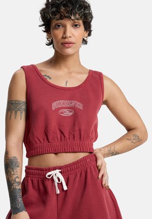 Kobieta z krótkimi kręconymi włosami, ubrana w czerwony krótki top Quiksilver i pasujące spodnie ze ściągaczem, pokazująca tatuaże na obu ramionach z neutralnym wyrazem twarzy.