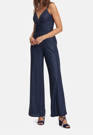 Kobieta ubrana w granatowy kombinezon bez rękawów z głębokim dekoltem w serek, szerokimi nogawkami i dopasowanymi granatowymi sandałami na obcasie.