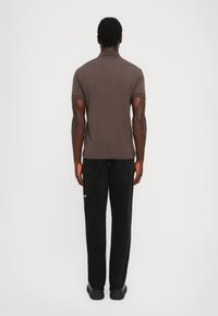 Mann som står med ryggen til, iført en brun kortermet polo skjorte og svarte jeans med svarte sko, mot en ensfarget hvit bakgrunn.