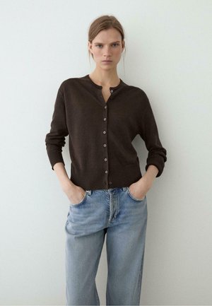 Donna con capelli biondi in piedi contro un muro bianco, indossa un cardigan marrone scuro abbottonato e jeans azzurri con le mani nelle tasche.