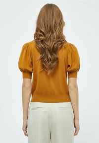 Senapsgul stickad tröja med puffärmar, ribbad nederkant och figursydd silhuett, i kombination med ljusbeiga byxor, sedd bakifrån.