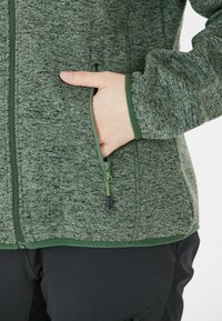 Grön texturerad jacka med dragkedja och en hand i en sidoficka, med mörkgrön kant och en slät dragkedjeöppnare. Svarta byxor syns nedanför.