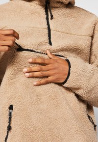 Pullover in pile beige con design a zip a un quarto, zip e rifiniture nere, caratterizzato da una superficie testurizzata e cappuccio regolabile con cordino.