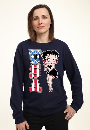 Sweatshirt marine avec un graphique représentant "USA" en rayures rouges et blanches, des étoiles bleues, et un personnage de dessin animé en robe noire tenant un microphone.