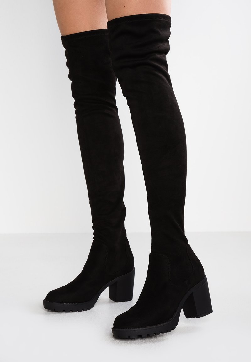Bottes noires en daim montant au-dessus du genou, dotées d'un talon carré épais, d'un bout arrondi et d'une tige texturée avec un design sans coutures et sans éléments métalliques visibles.