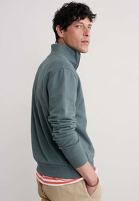 Grau-grüne Zip-Up-Jacke mit langen Ärmeln, gerippten Bündchen und Saum. Über einem weißen gestreiften Shirt getragen, kombiniert mit beigen Hosen.