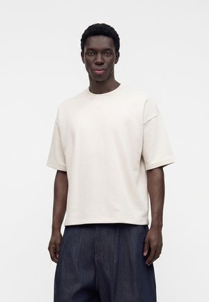 Uomo che indossa una camicia oversize tinta unita bianco sporco a maniche corte e pantaloni larghi blu scuro, in piedi su uno sfondo bianco.