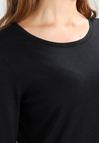 Chemise noire à manches longues avec un col arrondi, confectionnée en coton lisse, présentant une silhouette ajustée et sans motifs ou accents visibles.